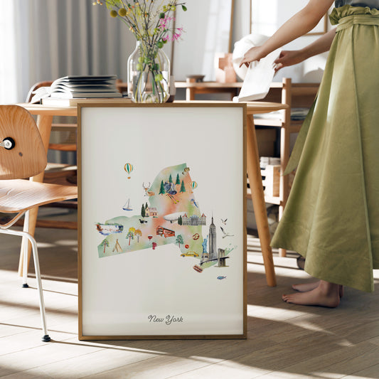 A framed watercolor illustration of New York is displayed on a table, with a person standing next to it.