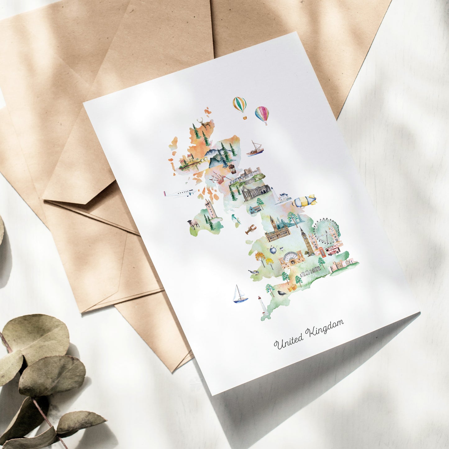 A card depicting various landmarks and scenes from the United Kingdom, including hot air balloons, a ferris wheel, and a castle, is displayed on a white surface alongside some envelopes.