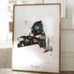 A framed collage of various New York-themed illustrations, including landmarks, buildings, and people, is displayed on a wooden floor.