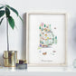 a framed watercolor illustration of Rhode Island, with various elements such as trees, buildings, and a hot air balloon. The illustration is displayed on a white shelf, accompanied by a potted plant and a small glass jar.