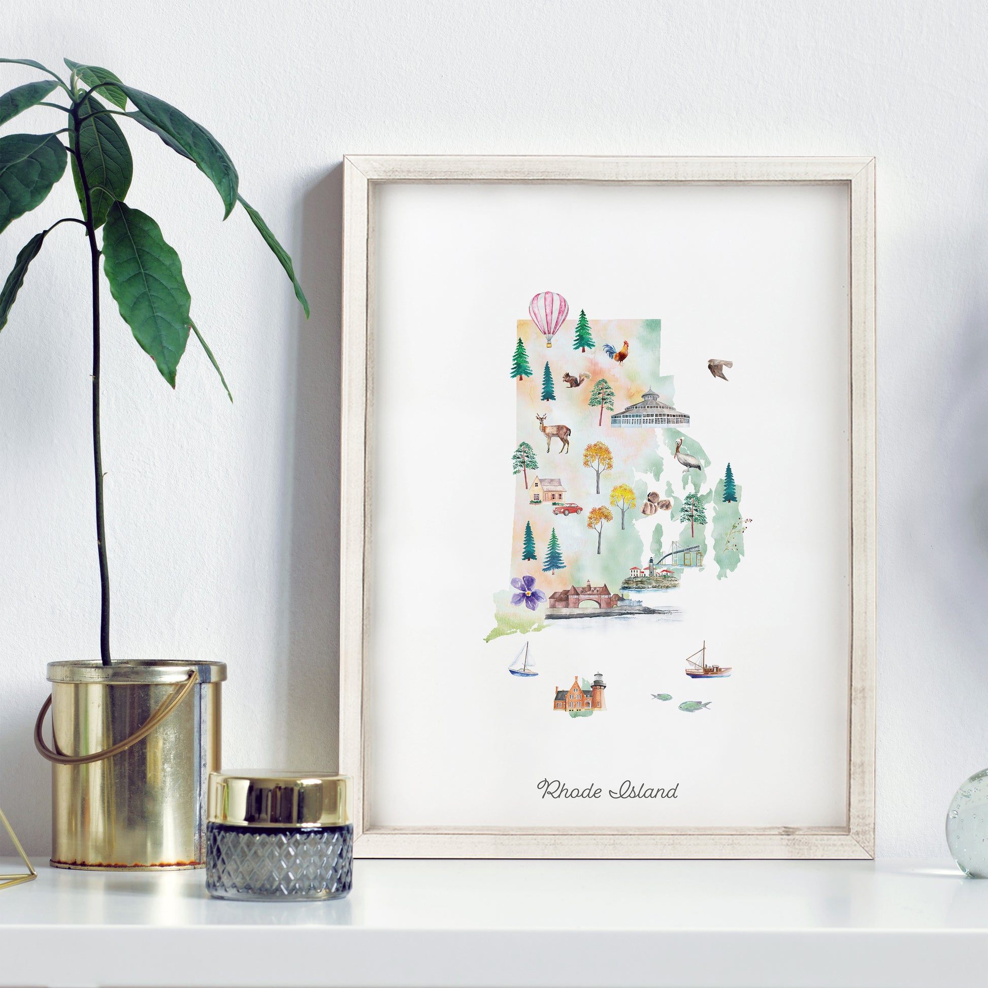 a framed watercolor illustration of Rhode Island, with various elements such as trees, buildings, and a hot air balloon. The illustration is displayed on a white shelf, accompanied by a potted plant and a small glass jar.