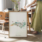 A framed watercolor illustration of North Carolina, featuring various landmarks and scenes, is displayed on a table in a room with a person standing nearby.