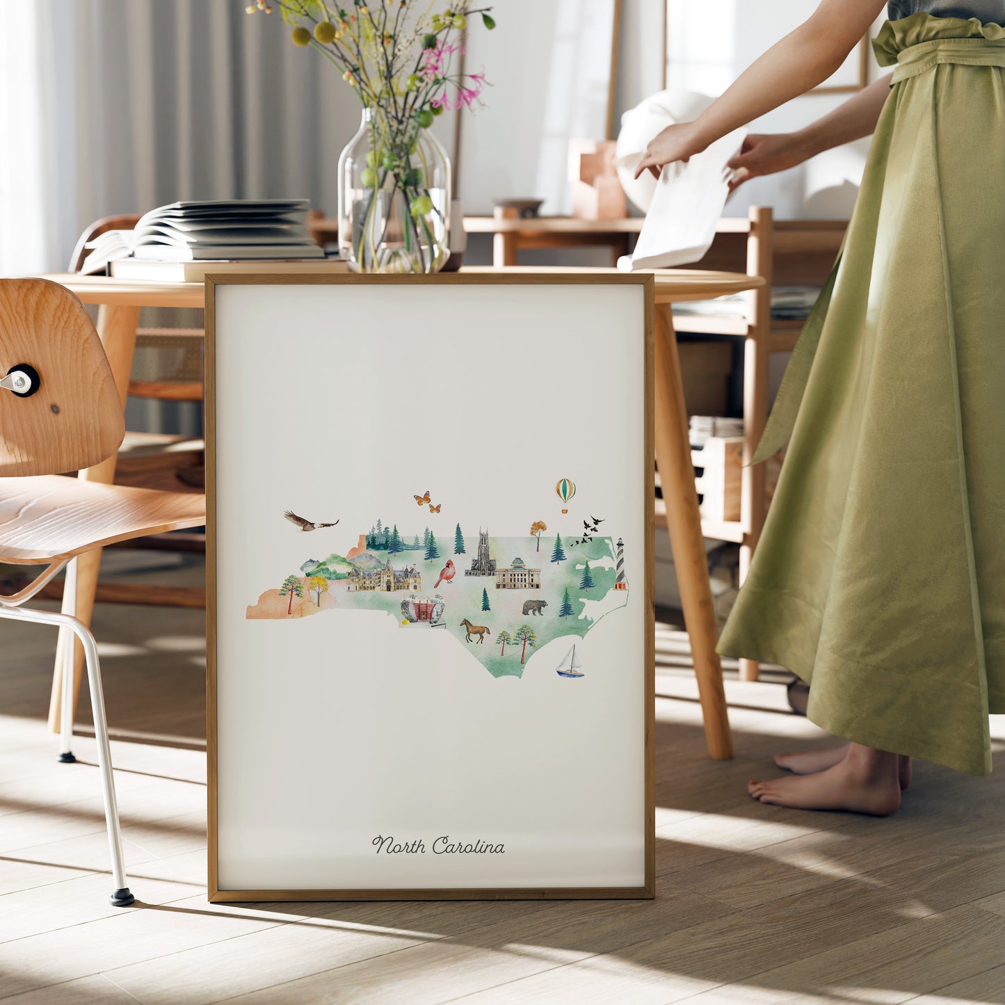 A framed watercolor illustration of North Carolina, featuring various landmarks and scenes, is displayed on a table in a room with a person standing nearby.