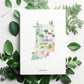 The image is a watercolor illustration of the state of Indiana, surrounded by green leaves and flowers.