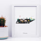 A framed illustration of Tennessee's state map, featuring various landmarks and symbols, is displayed next to a potted cactus plant.