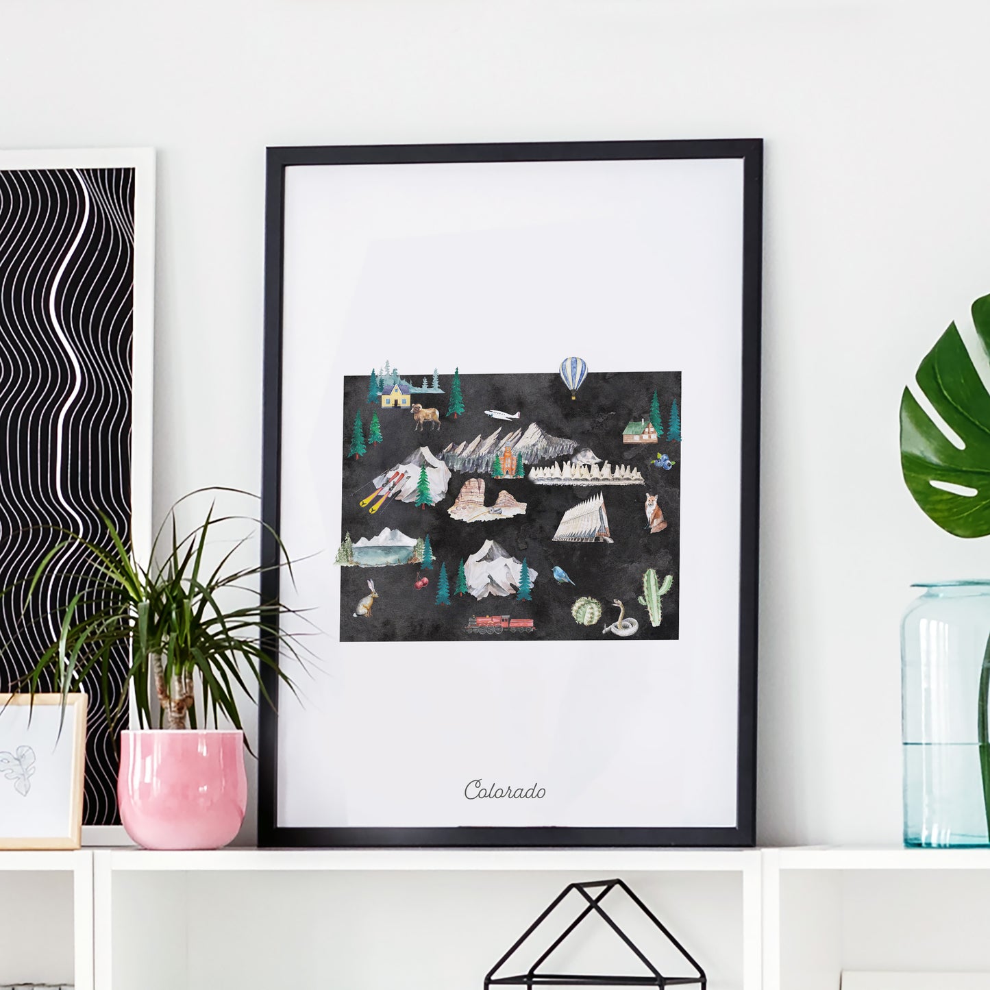 a framed artwork depicting various landscapes and elements of Colorado, including mountains, forests, and a hot air balloon, displayed on a shelf.
