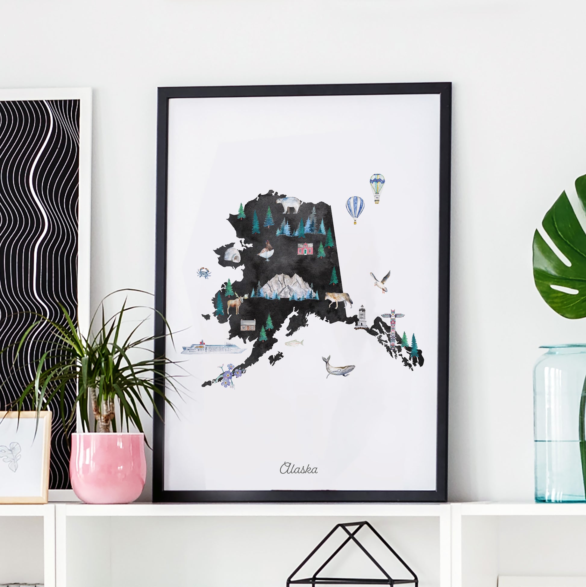 a framed illustration of Alaska, with various elements such as mountains, forests, and hot air balloons depicted on the map.