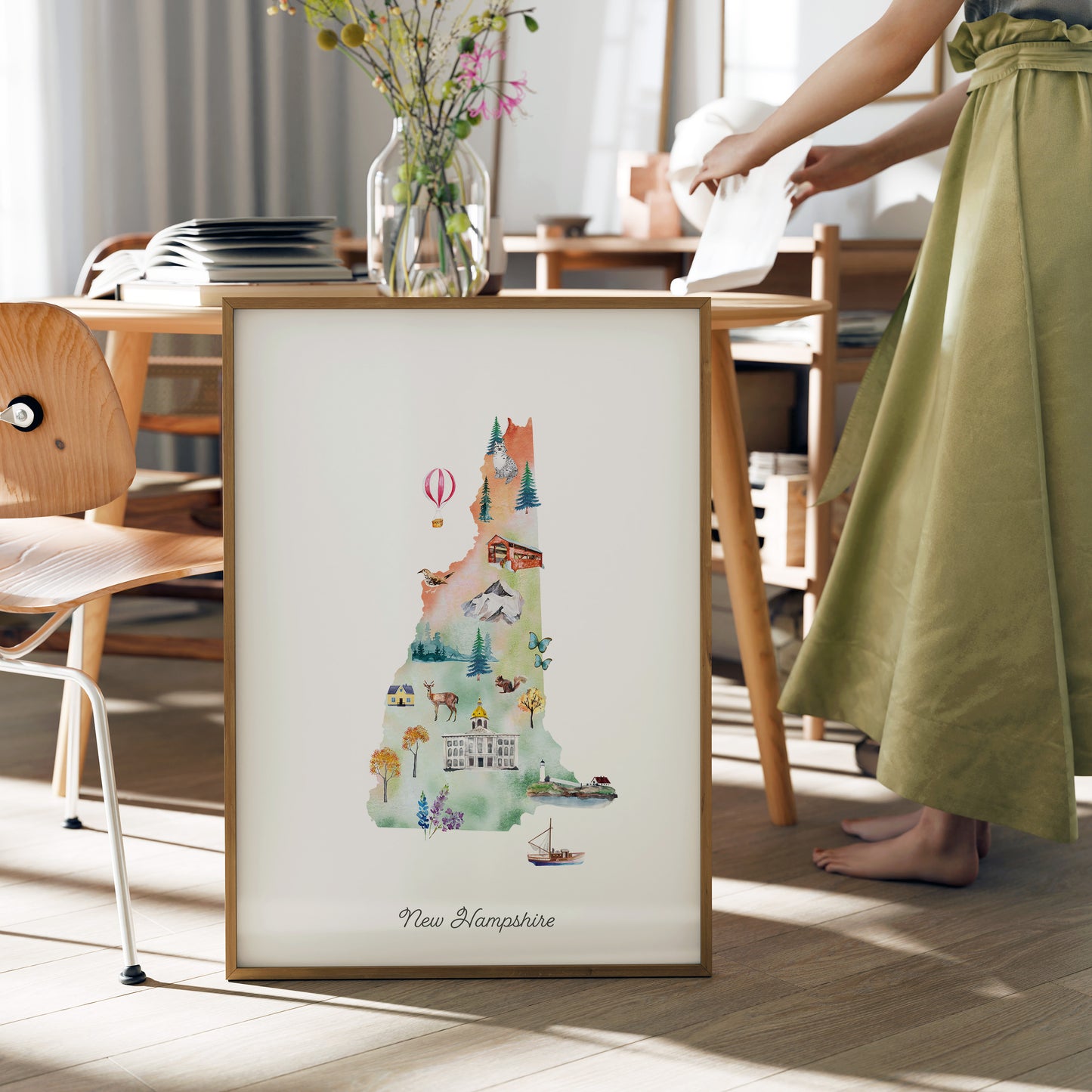 A framed watercolor illustration of New Hampshire, featuring various landmarks and scenes, is displayed on a table in a room with a person standing nearby.