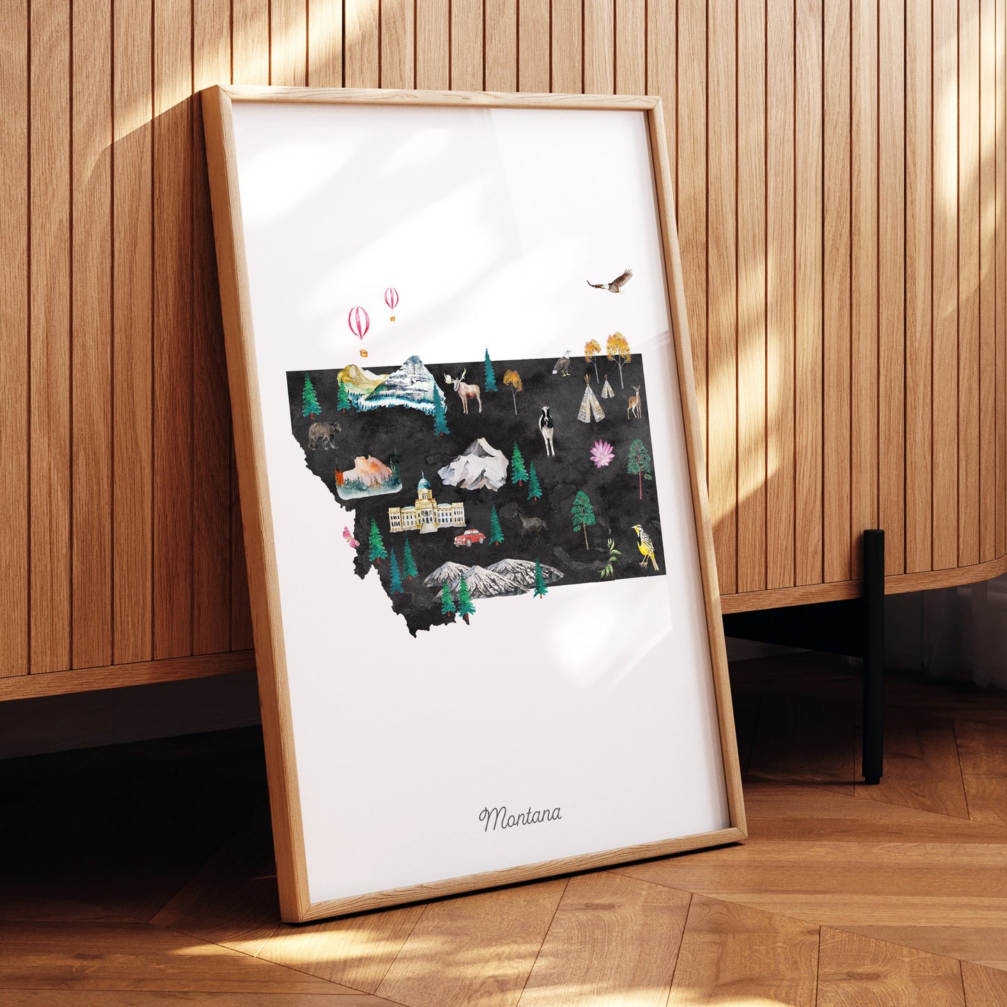 A framed illustration of Montana, featuring various landmarks and natural scenery, is displayed on a wooden surface.