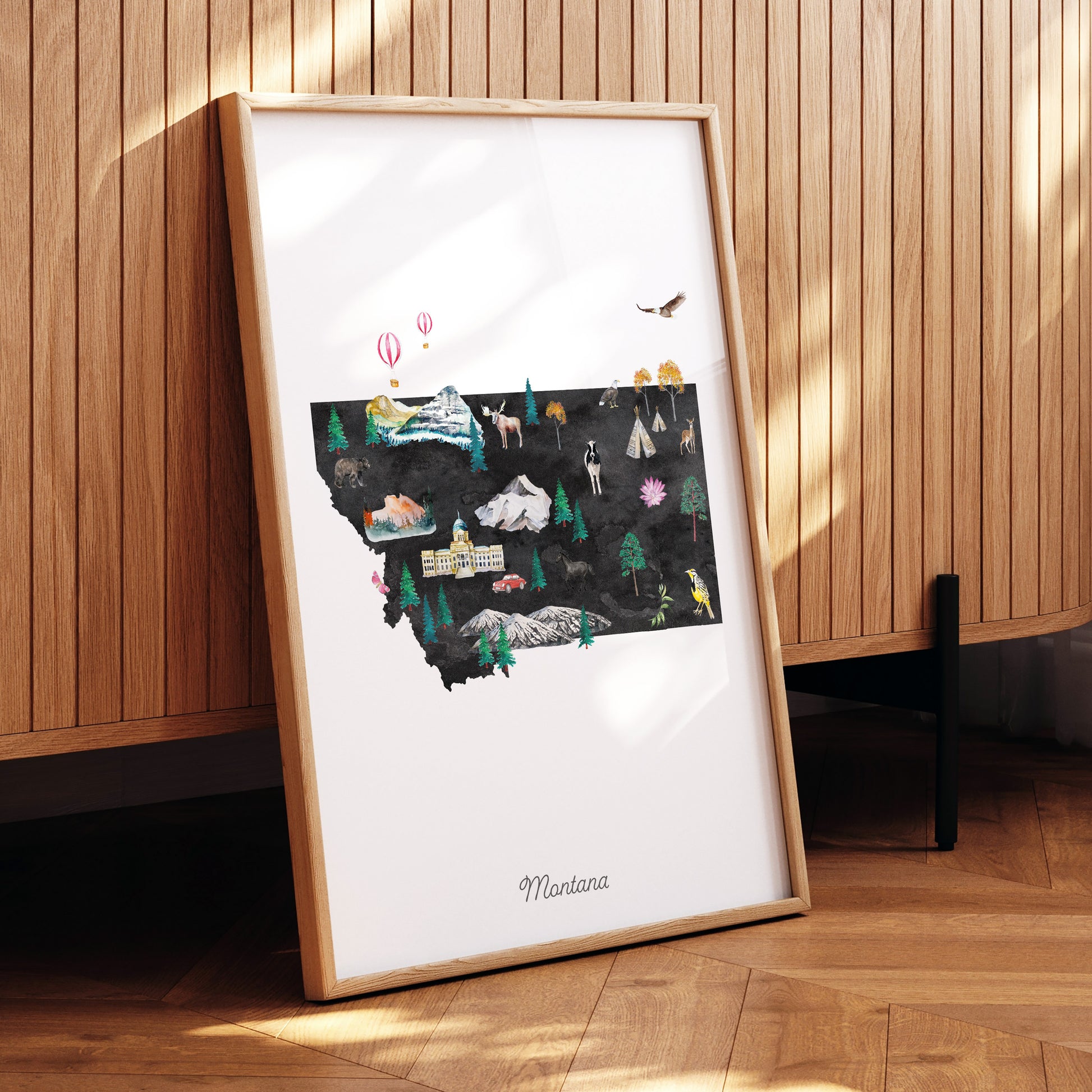A framed illustration of Montana, featuring various landmarks and natural scenery, is displayed on a wooden surface.