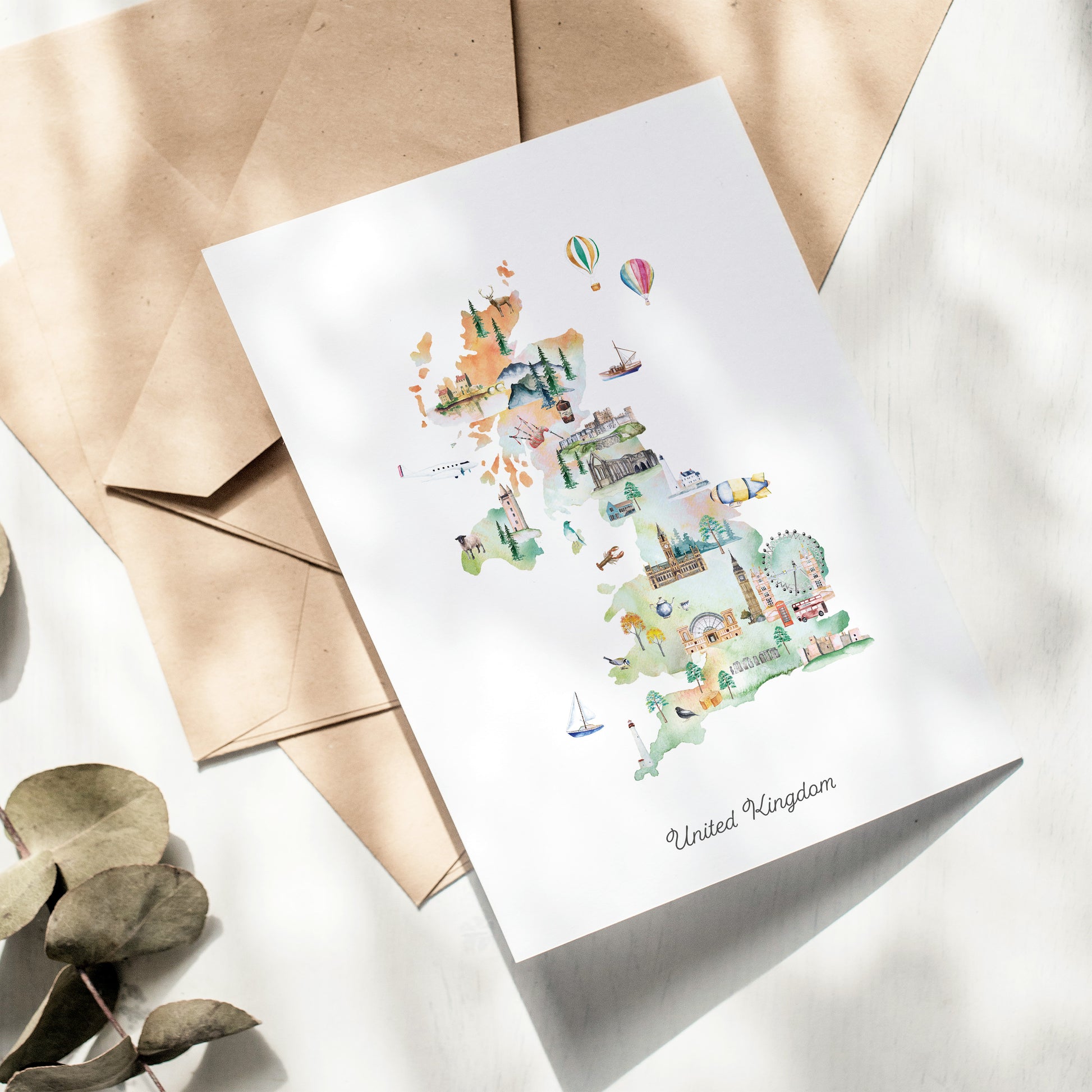 A card depicting various landmarks and scenes from the United Kingdom, including hot air balloons, a ferris wheel, and a castle, is displayed on a white surface alongside some envelopes.