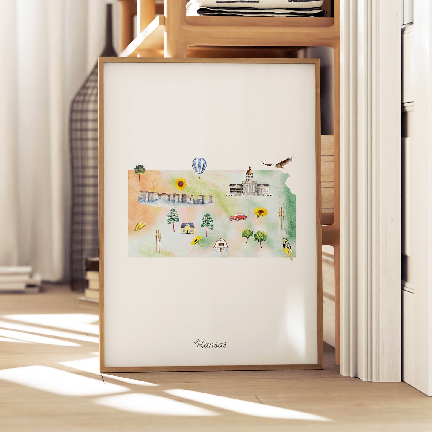 A framed watercolor painting depicting various scenes and landmarks of Kansas, including a hot air balloon, a castle, and a field with trees.