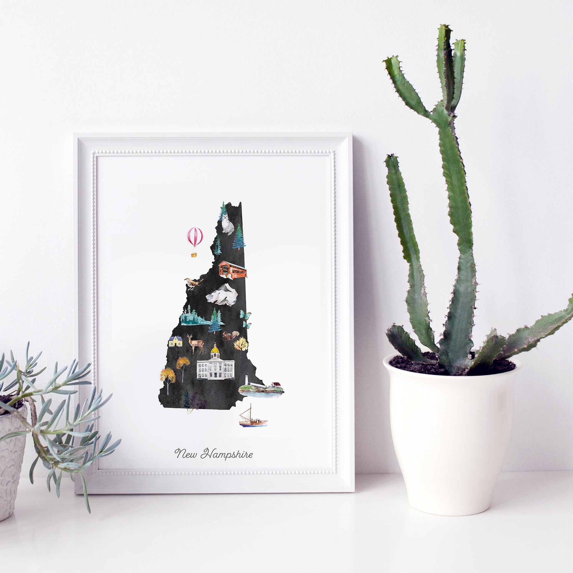 a framed illustration of the state of New Hampshire, with various landmarks and symbols representing the state's unique features. Next to the framed illustration, there is a potted plant and a white vase, creating a decorative and harmonious display.