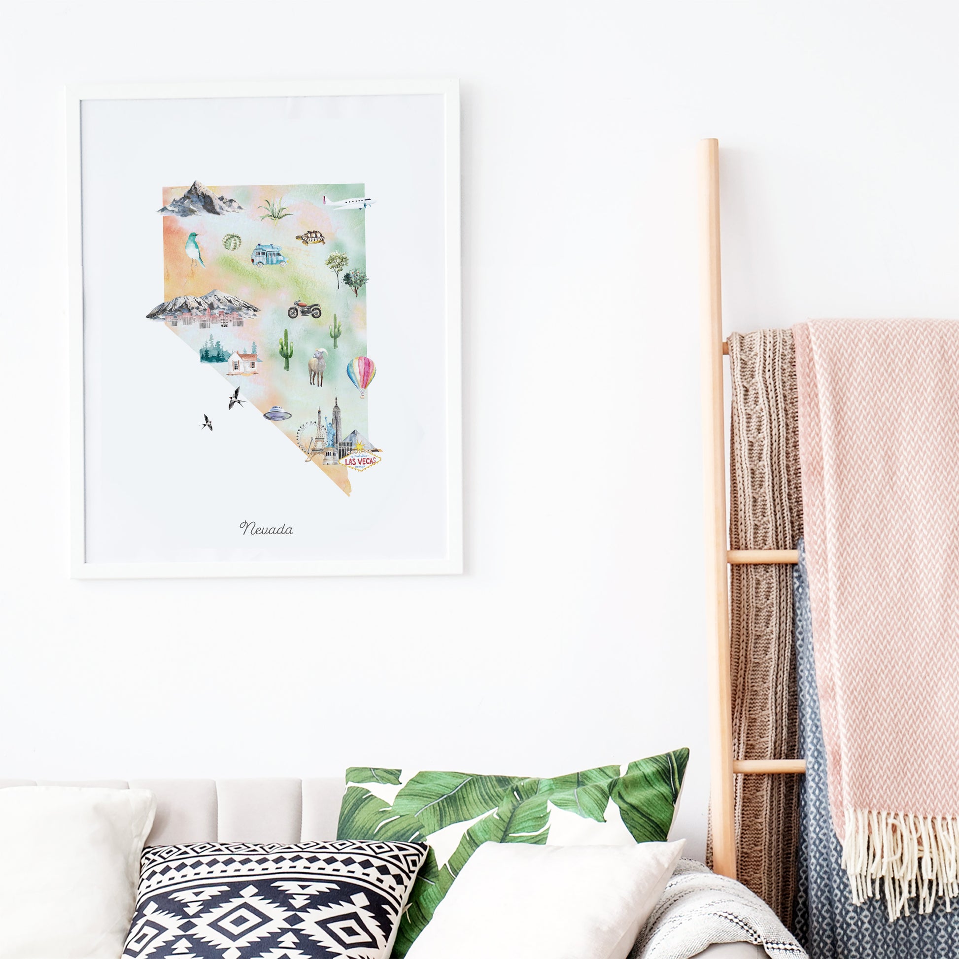 a framed watercolor painting of a map of Nevada, with various illustrations of landmarks and symbols, hanging on a white wall above a couch with patterned pillows.
