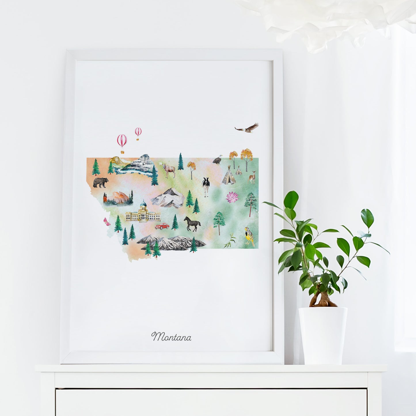 a framed watercolor painting of Montana, with various illustrations of landmarks, animals, and natural elements. The painting is displayed on a white shelf, and there is a potted plant next to it.