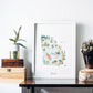 a framed illustration of Georgia, with various landmarks and natural elements such as trees and mountains. The illustration is displayed on a white wall, and there are some decorative items placed around it, including a potted plant, a wooden crate, and a glass vase.