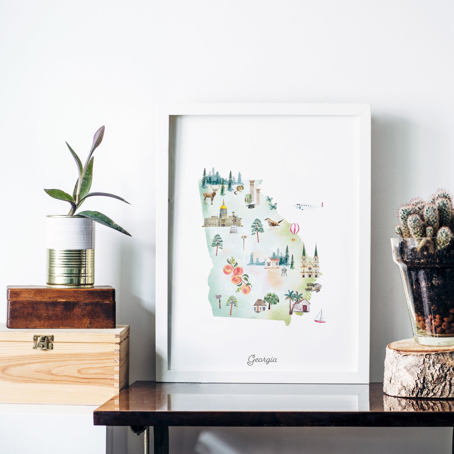 a framed illustration of Georgia, with various landmarks and natural elements such as trees and mountains. The illustration is displayed on a white wall, and there are some decorative items placed around it, including a potted plant, a wooden crate, and a glass vase.