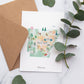 a greeting card with a watercolor illustration of various landmarks and scenes from Arkansas, including a hot air balloon, a deer, a building, and a tree. The card is placed on a marble surface, with a brown envelope and some greenery nearby.