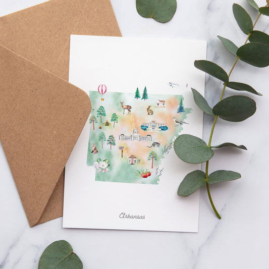 a greeting card with a watercolor illustration of various landmarks and scenes from Arkansas, including a hot air balloon, a deer, a building, and a tree. The card is placed on a marble surface, with a brown envelope and some greenery nearby.