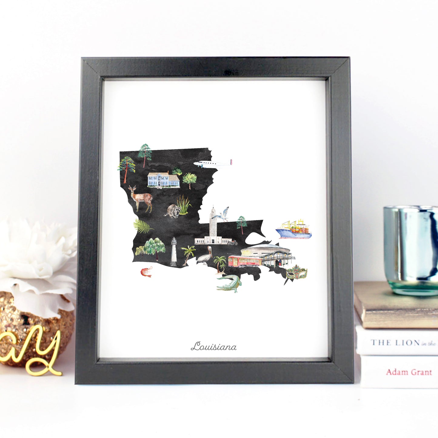 A framed illustration of Louisiana, featuring various landmarks and elements of the state, such as a lighthouse, boats, and buildings, is displayed on a shelf next to a stack of books and a decorative item.
