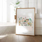 A framed watercolor painting depicting various scenes and landmarks of Pennsylvania, including a deer, a hot air balloon, and a building, is displayed on a wooden floor in a room with a potted plant in the background.