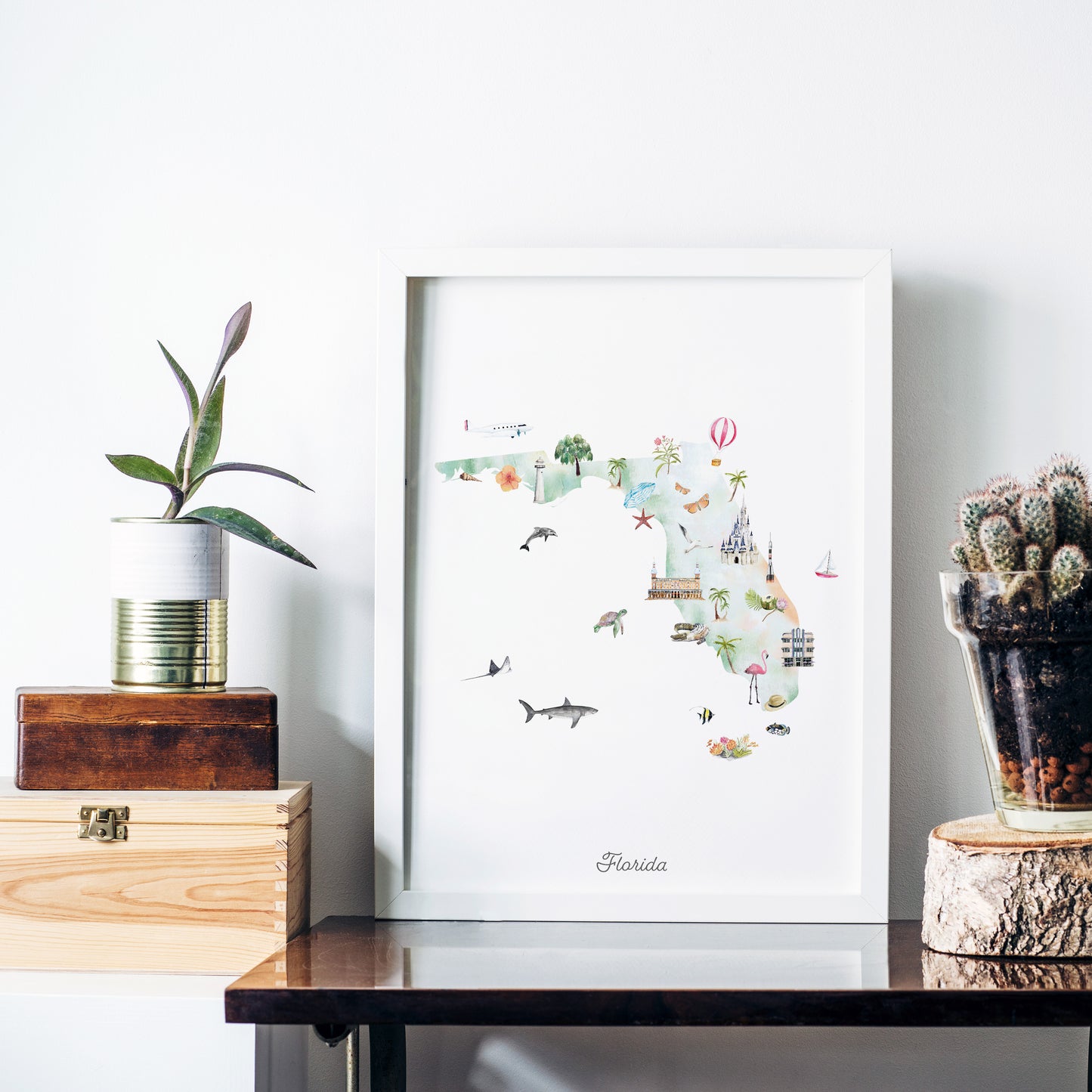 a framed map of Florida, with various illustrations of landmarks and attractions, such as palm trees, cacti, and hot air balloons, all set against a white background. The map is displayed on a wooden shelf, with a potted plant and a glass vase nearby.