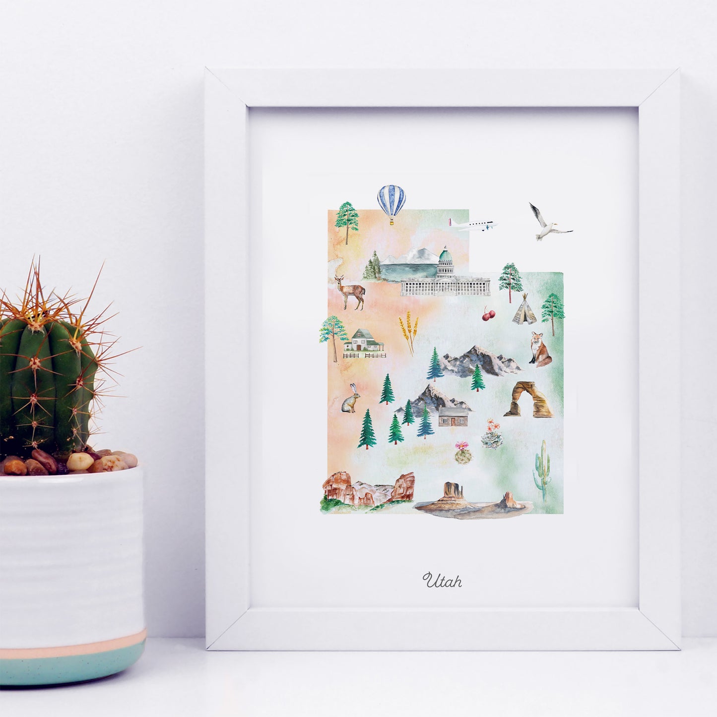 A framed watercolor illustration depicting various scenes and landmarks of Utah, including mountains, cacti, buildings, and wildlife.