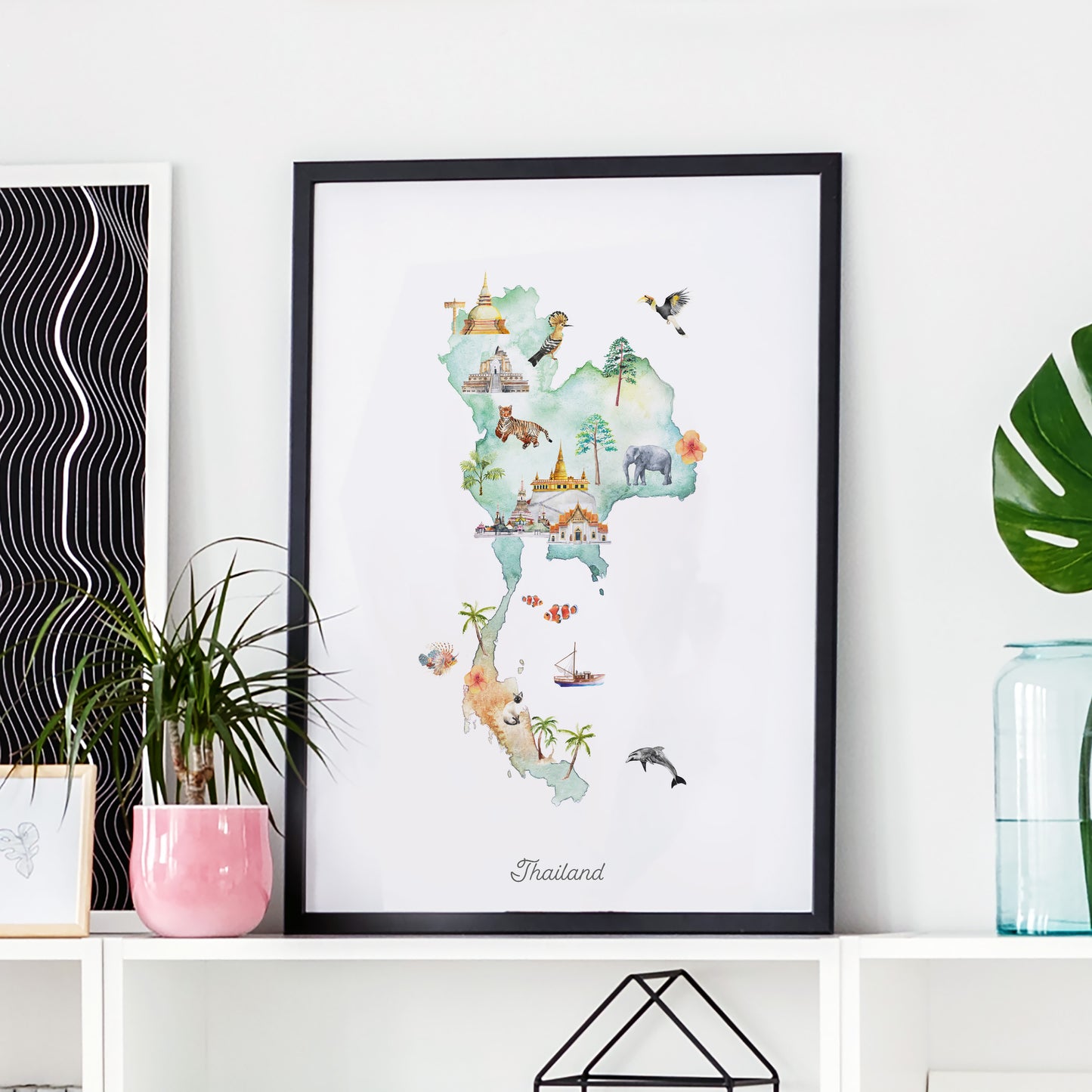 A framed watercolor map of Thailand, featuring various landmarks and animals, is displayed on a shelf alongside a pink vase and a green plant.