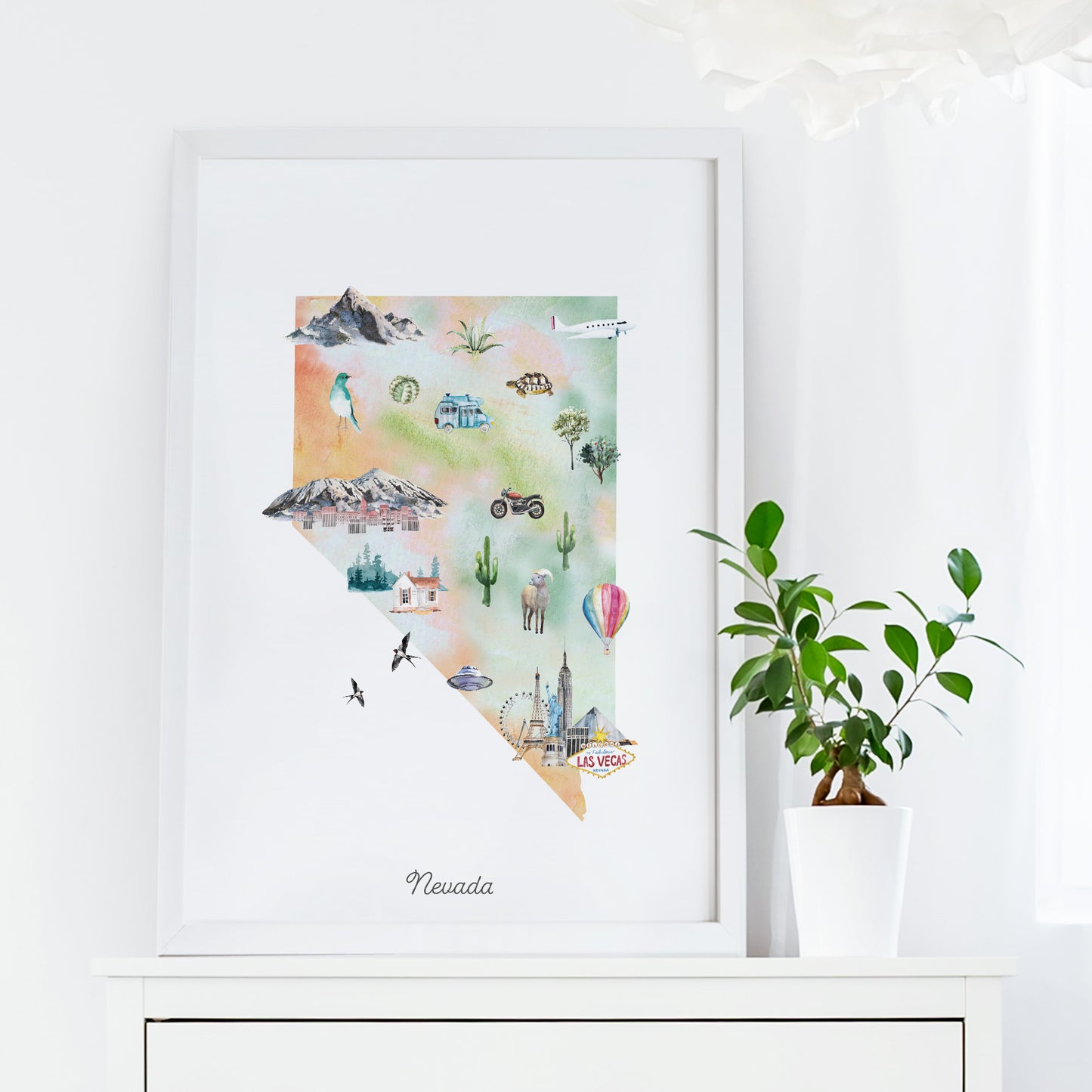 a framed watercolor painting of the state of Nevada, with various landmarks and symbols such as mountains, cacti, and hot air balloons. The painting is displayed on a white shelf next to a potted plant.